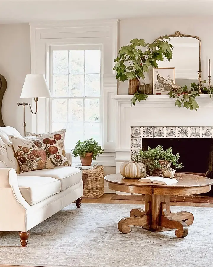 A cozy living room with a white sofa, floral pillow, round wooden coffee table with pumpkin, fireplace adorned with greenery, and a large mirror above the mantel creates inviting farmhouse fall decor.