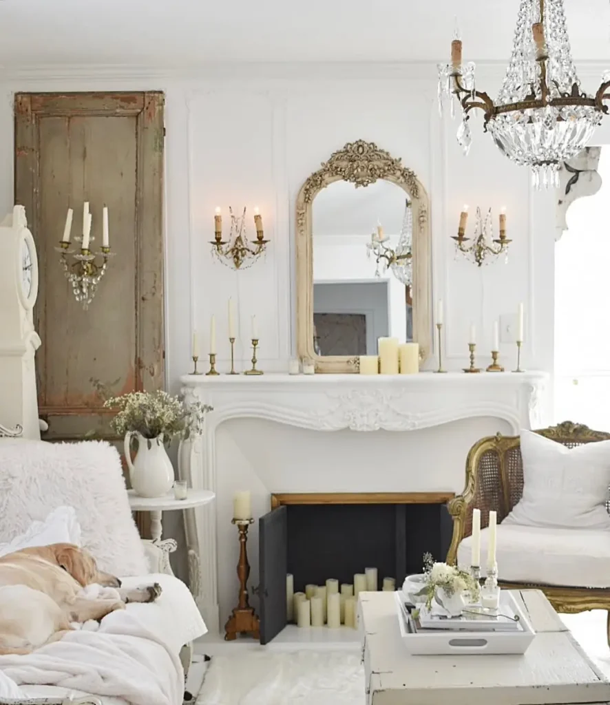 Elegant living room with a white fireplace, ornate mirror, candles, vintage chandelier, and a dog sleeping on a white sofa. Neutral tones and antique furnishings bring timeless French country decor to the space.