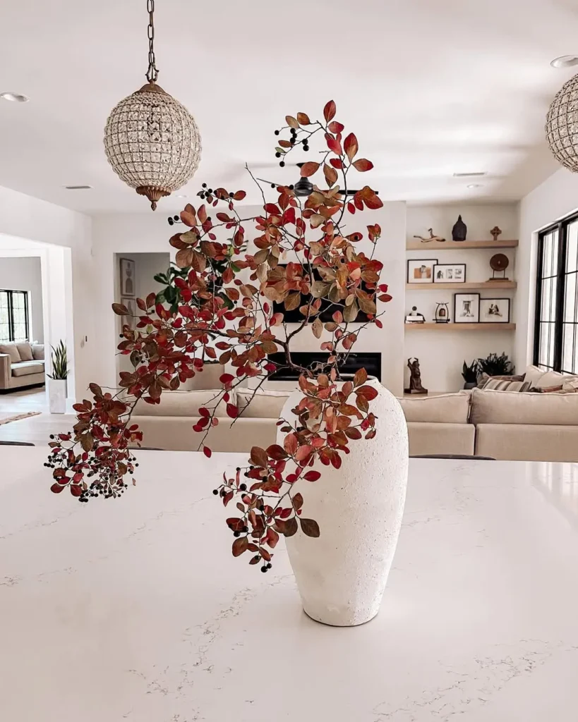 A white vase with branches of red and brown leaves sits on a marble countertop in a modern, well-lit living room—perfect inspiration from a modern fall decor guide.