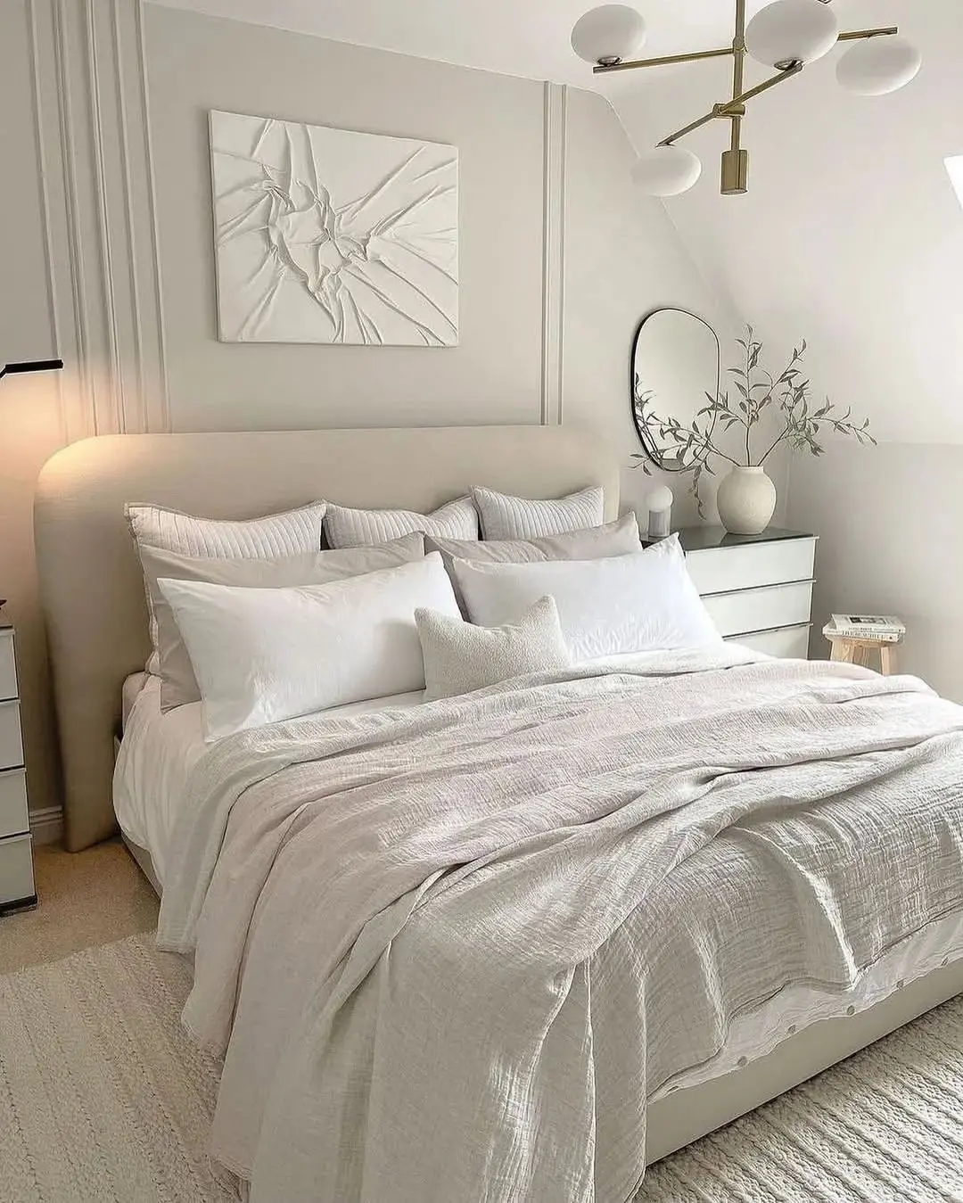 A neatly made bed with white and beige bedding sits in a minimalist bedroom with light walls, modern decor, and a contemporary light fixture overhead.