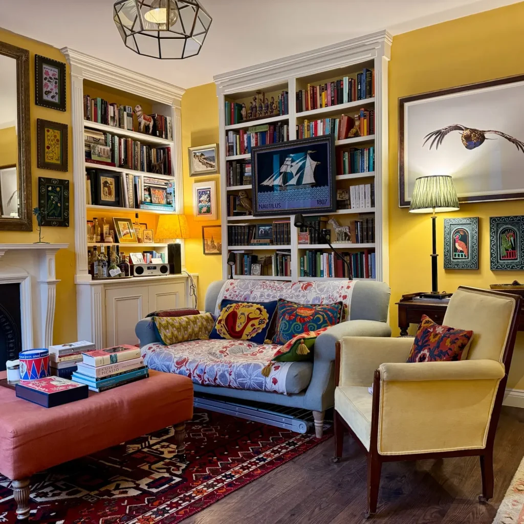 A cozy living room with yellow walls, bookshelves filled with books, a patterned sofa and armchair adorned with maximalist pillows, a red ottoman, framed art, and a floor lamp.