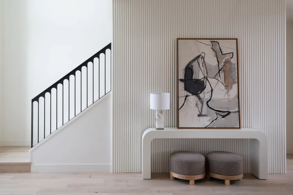A modern entryway features a sleek white console table, two round gray ottomans, a contemporary table lamp, and abstract framed artwork set against a ribbed wall beside a staircase with a striking black railing.