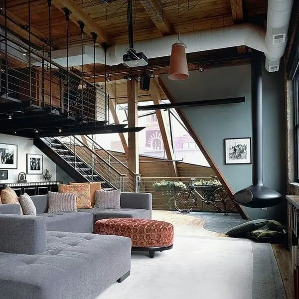 Modern industrial living room in a spacious loft featuring exposed wood beams, black metal staircase railing, concrete floors, oversized gray sectional sofa, and industrial fireplace in an open-concept urban interior.