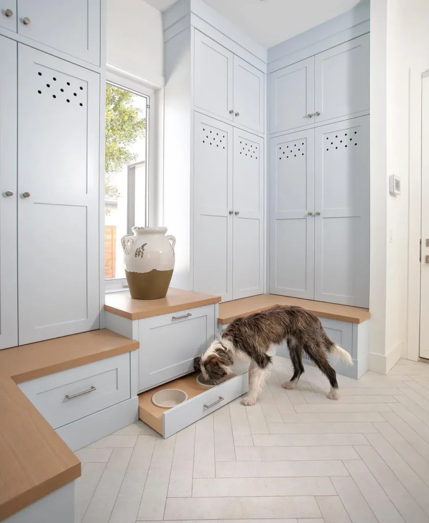 Built-in mudroom pet feeding station with pull-out dog bowls, light blue cabinetry, wood bench seating, and herringbone tile flooring for a functional, pet-friendly entryway.