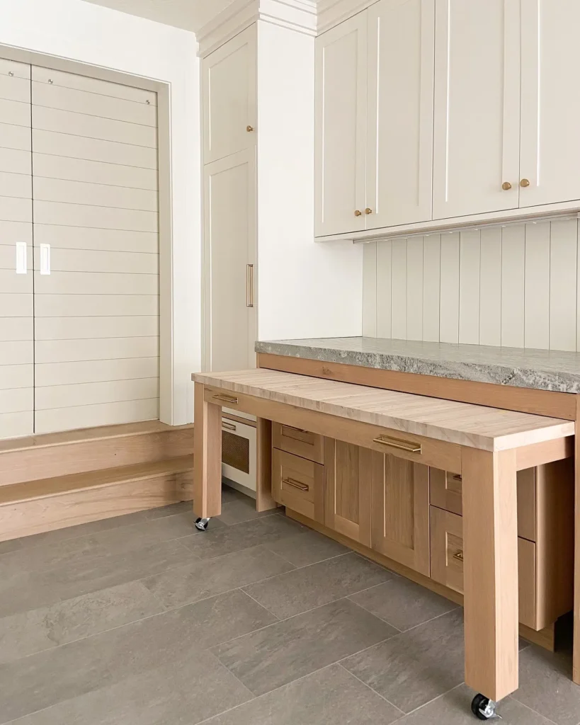 Built-in laundry room with a pull-out wooden folding station on casters, shaker-style cabinets, stone countertop, beadboard backsplash, and a clean, functional custom laundry workspace.